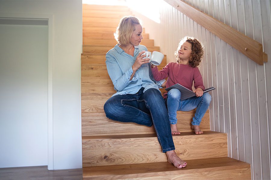 About Our Agency - Portrait of Mother and Daughter Sitting on a Wooden Staircase at Home While They Enjoy a Cup of Tea